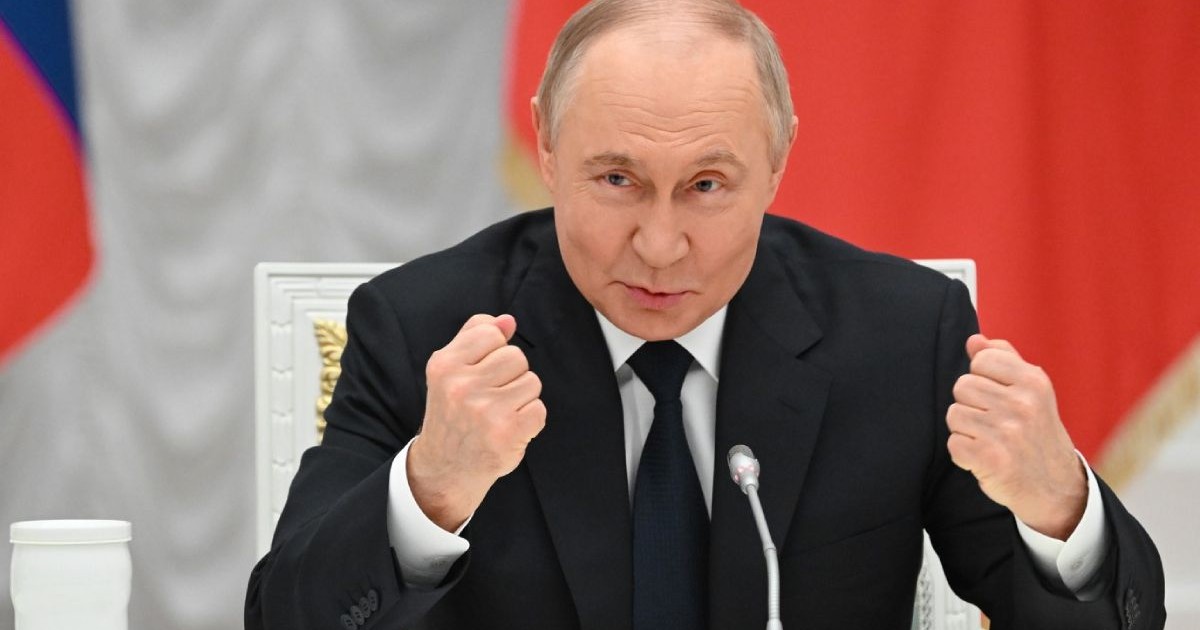 În imagine liderul rus Vladimir Putin, cu mâinile ridicate și pumnii strânși, stând la masă, zâmbind, pe un fundal roșu
