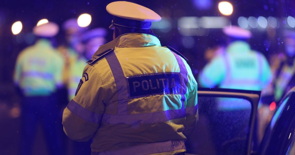Polițist îmbrăcat în uniformă, fotografiat din spate, noaptea, lângă o mașină de Poliție cu ușa deschisă
