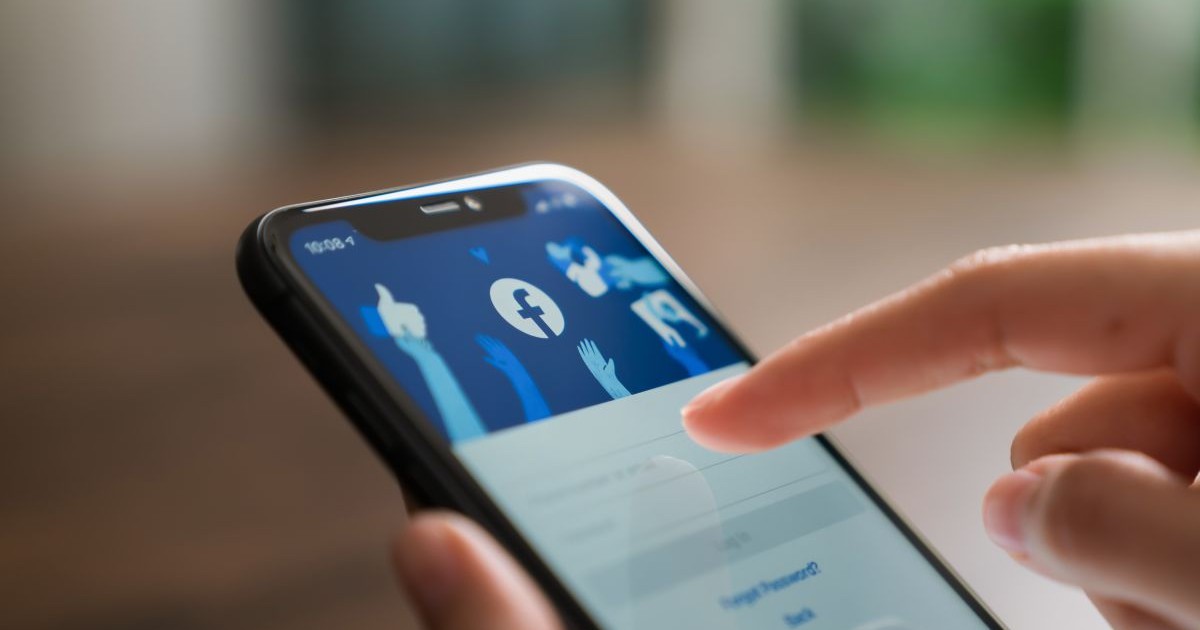 Telefon mobil cu aplicația Facebook deschisă pe display, ținut în mână de o persoană, cu fundal blurat