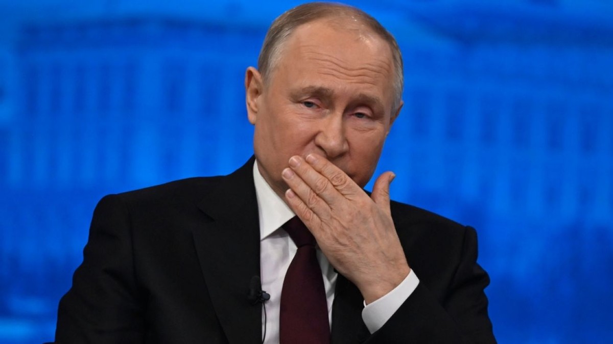 Vladimir Putin, îmbrăcat în costum negru, cu cămașă albă și cravată albastră, cu mâna la gură, pe fundal de culoare albastră