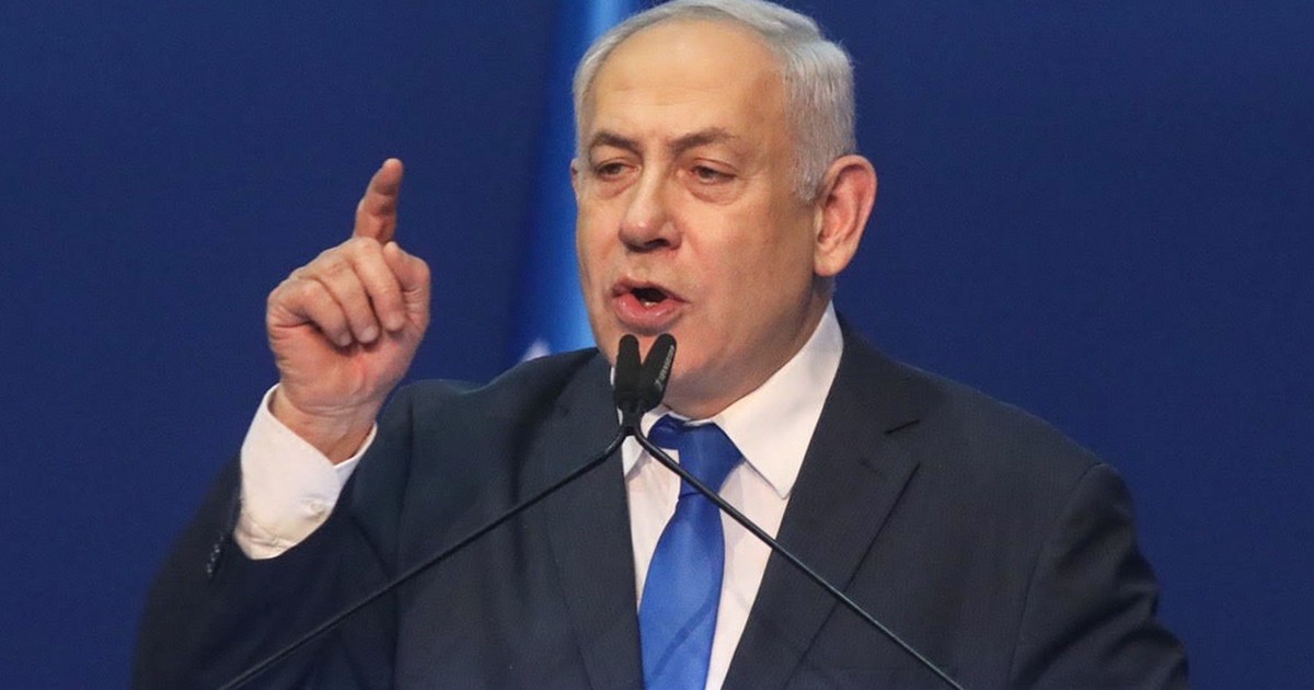 Benjamin Netanyahu, susține un discurs la microfon, purtând un costum bleumarin și o cravată albastră, ținând mâna dreaptă ridicată, cu degetul arătător ridicat