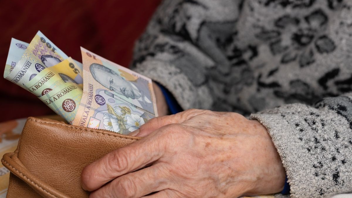 O bătrână cu pulover pe ea scoate dintr-un portofel maro, pe care îl ține cu o mână, câteva bancnote românești