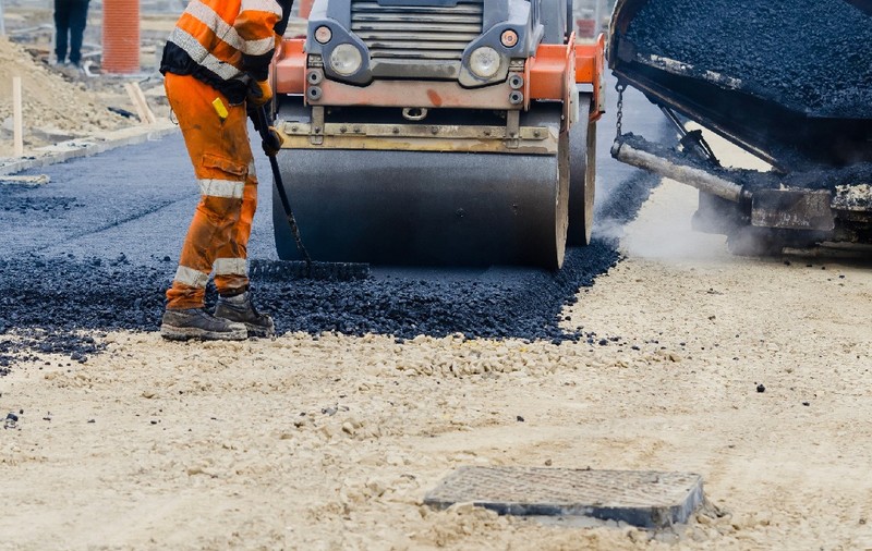Lucrătorii din construcții în echipamente de înaltă vizibilitate aplică asfalt proaspăt fierbinte pe suprafața drumului