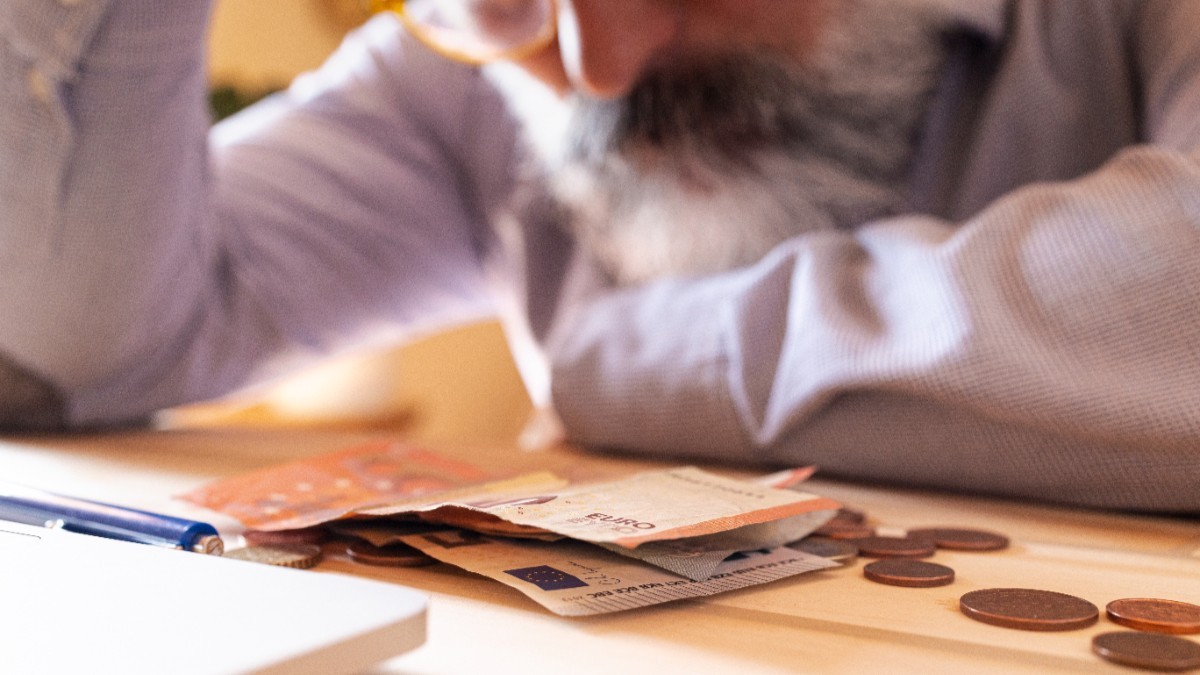 Un bărbat cu ochelari care stă la birou, cu mâna dreaptă pe frunte. Pe birou se află mai multe bancnote și monede de euro 