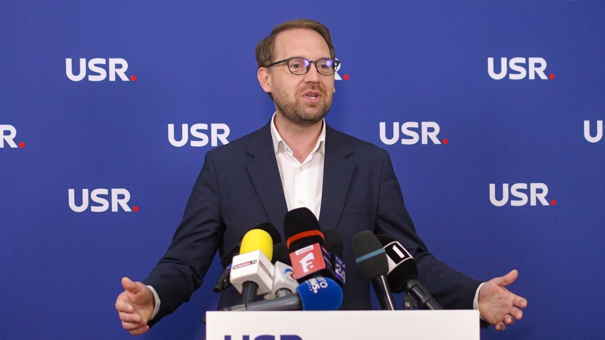 Dominic Fritz îmbrăcat într-un sacou negru și o cămașă albă, vorbind în fața unui pupitru cu sigla USR pe care se află mai multe microfoane, cu mâinile larg deschise