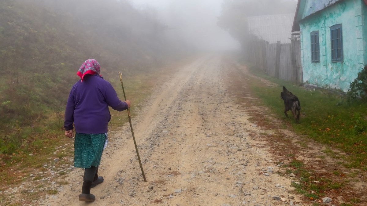 Femeie în vârstă, care se sprijină în baston, merge pe o uliță a unui sat din România, pe timp de ceață, cu un câine negru lângă ea, iar pe partea dreaptă o casă cu pereții albaștri-bleu