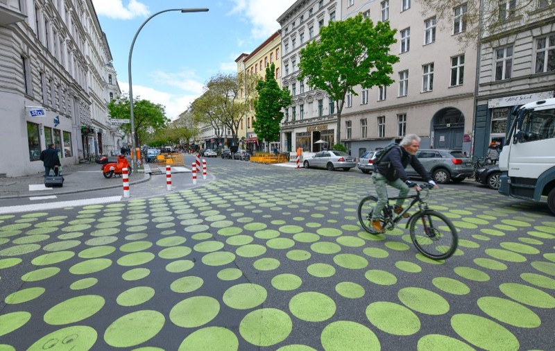 O stradă urbană mărginită de clădiri și copaci, unde carosabilul este pictat cu un model din cercuri galbene de diferite dimensiuni, pe care se deplasează un biciclist.   