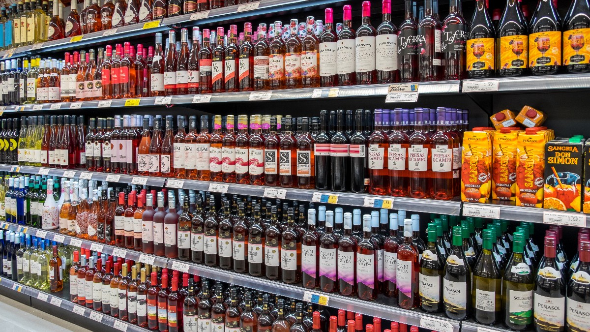 Imensa varietate de sticle de băuturi alcoolice, în principal vinuri roșii și roze, este expusă pe mai multe rafturi lungi, în secțiunea unui supermarket. Rafturile sunt pline ochi cu produse, sugerând o ofertă largă de etichete și sortimente pentru clienți.
