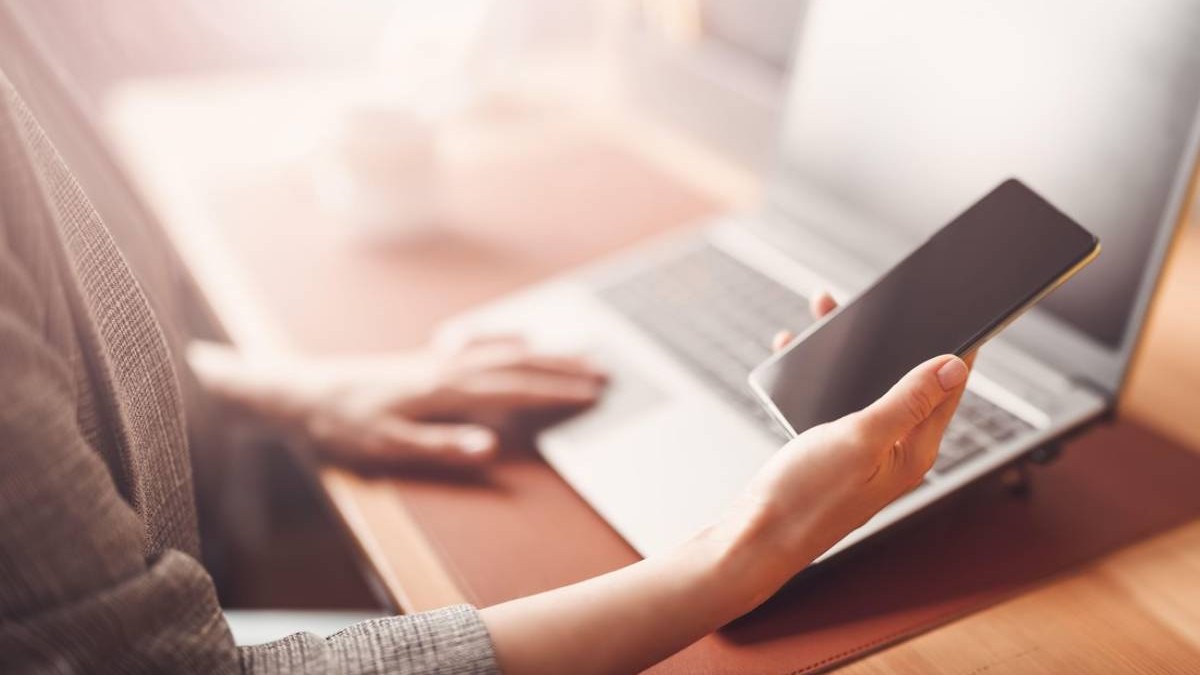 Femeie cu smartphone în mână lucrând la laptop la birou.