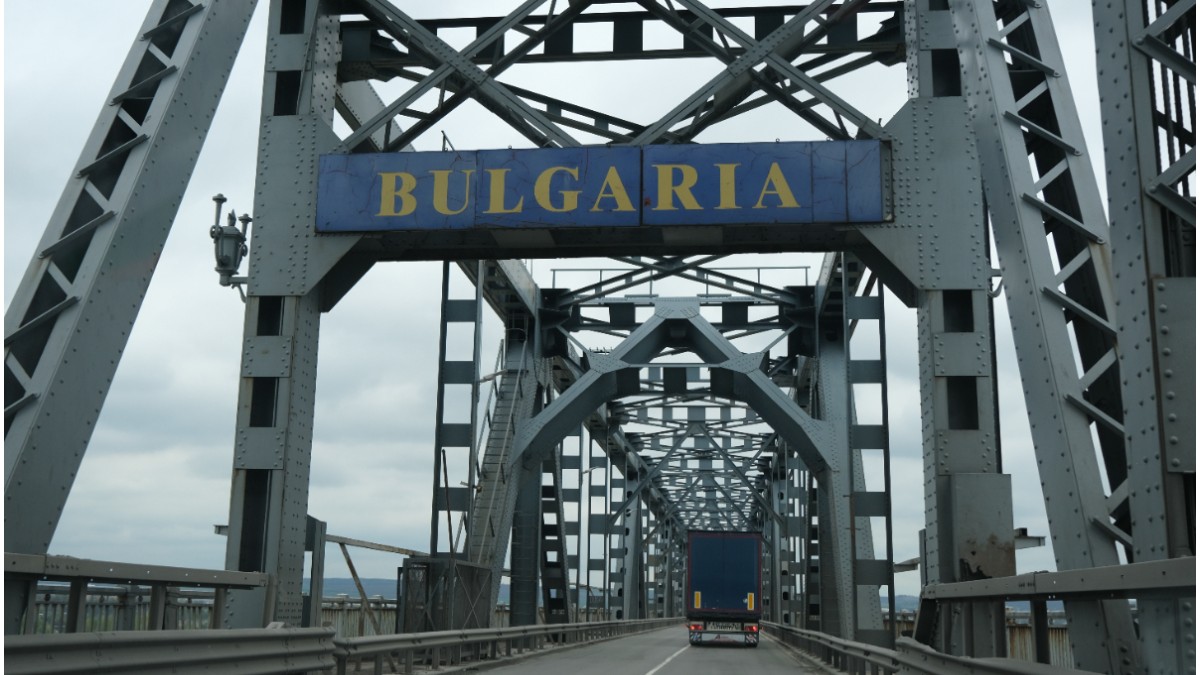 În imagine este un pod masiv cu structură metalică, pe care circulă un camion și deasupra căruia este montat un indicator albastru cu textul „BULGARIA”.