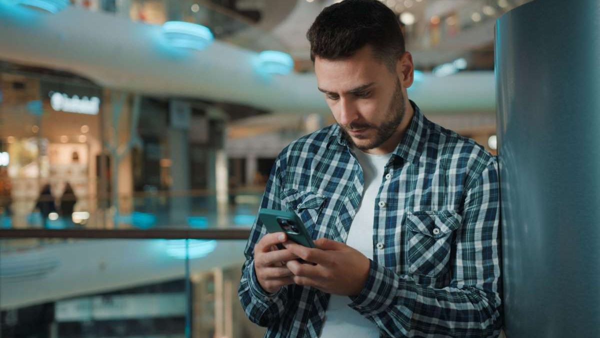 Un bărbat se uită concentrat la telefon în timp ce se află într-o clădire de sticlă.