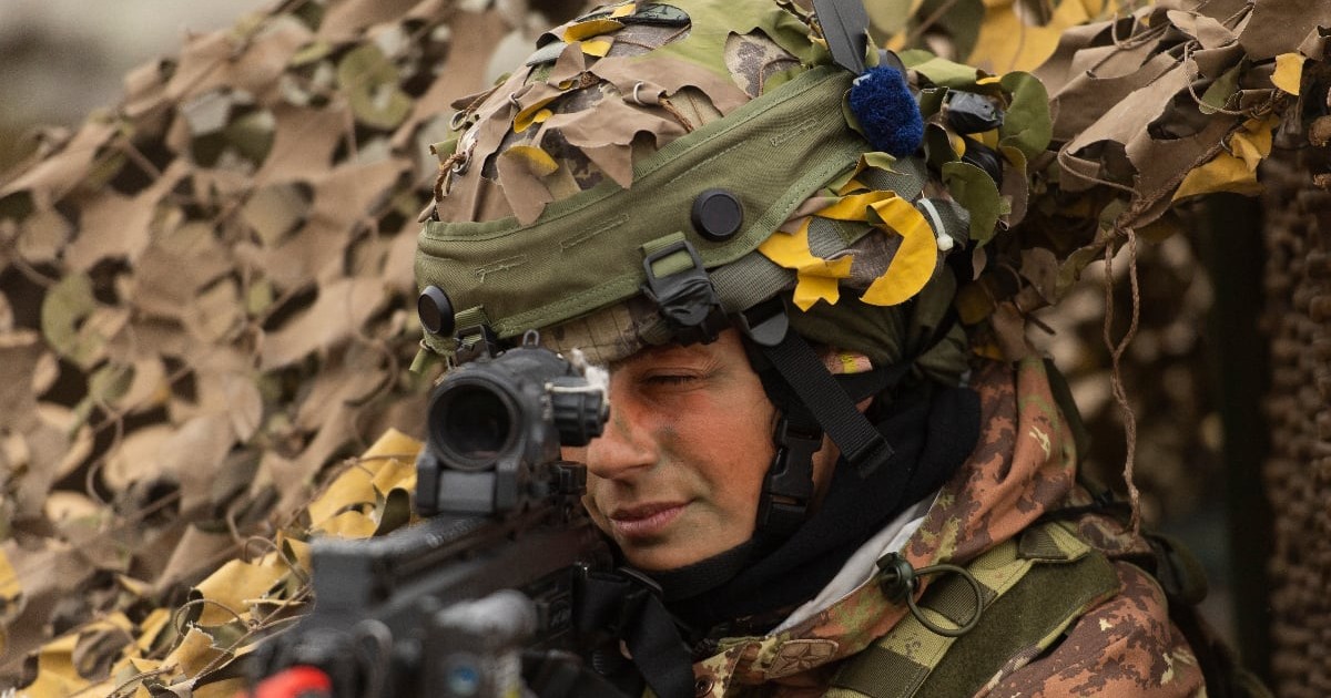 Un soldat al armatei italiene, echipat în uniformă de camuflaj și cască militară, țintește cu arma în timpul unui exercițiu. Pe uniforma sa este vizibil ecusonul cu drapelul Italiei, în timp ce în jur se află echipamente de camuflaj folosite pentru operațiuni militare.