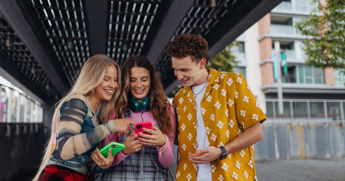 Imaginea surprinde trei tineri care privesc zâmbind ecranul unui telefon mobil, într-un spațiu urban modern, sub o structură metalică. Atmosfera este relaxată și sugerează socializare, apropiere și interes comun pentru conținutul digital.