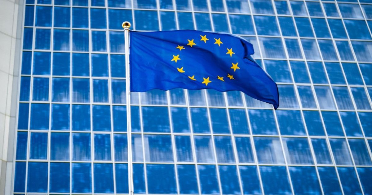 Imagine creativă cu steagul Uniunii Europene arborat în fața unei clădiri administrative din Bruxelles cu geamuri de culoare albastră