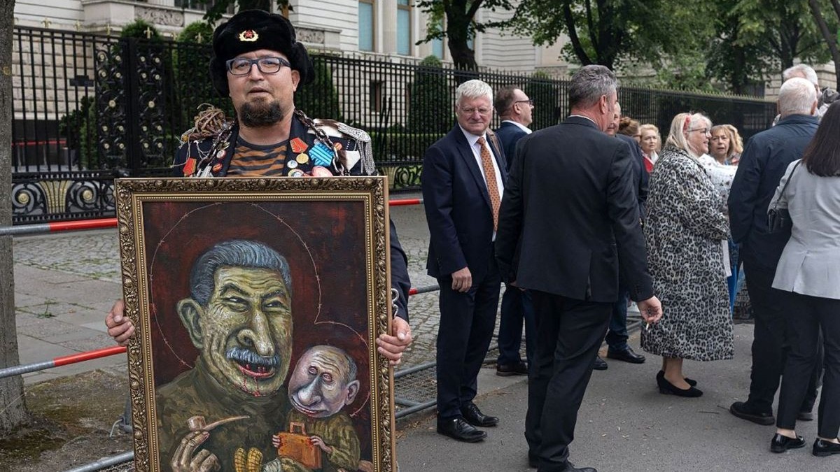Artistul rus Semion Skrepețki cu o căciulă „rusească” pe cap, ține în mâini un tablou în care Iosif Stalin îl ține în brațe pe „micuțul” Vladimir Putin, Berlin, Germania, 1 iunie 2026