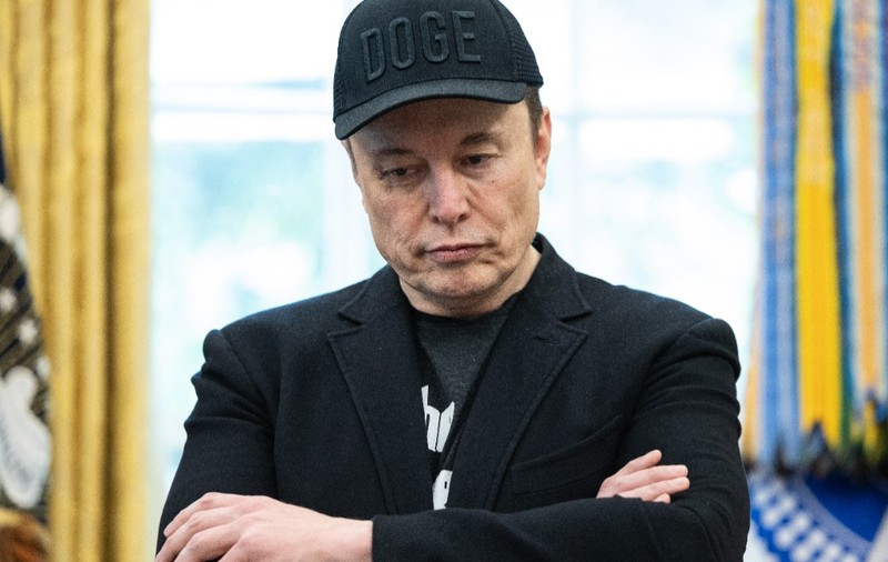 Elon Musk stând cu brațele încrucișate și privind în jos, îmbrăcat într-o jachetă neagră și purtând o șapcă neagră inscripționată cu textul „DOGE”.
