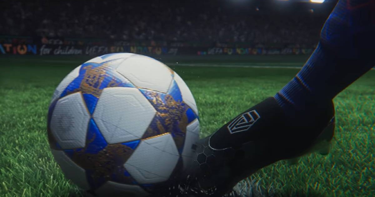 În imagine se vede un picior de jucător care lovește o minge de fotbal aflată pe gazonul unui stadion