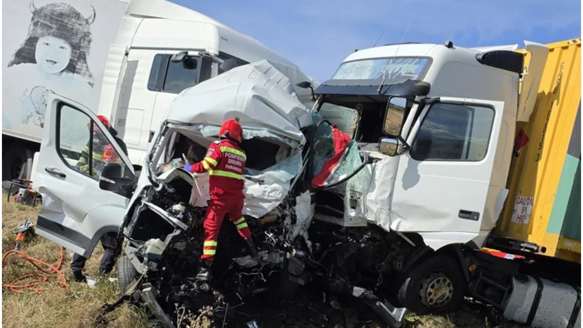 Imaginea prezintă urmările unui accident grav, în care o dubă albă este zdrobită de două TIR-uri, în timp ce un echipaj de pompieri și paramedici SMURD intervine la fața locului.