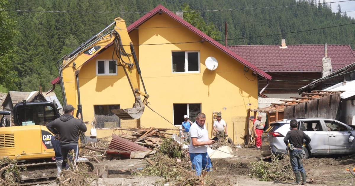 Pompierii și voluntarii de la Broșteni scot nămolul și lemnele din curtea unei case mari, galbene, avariată în timpul inundațiilor