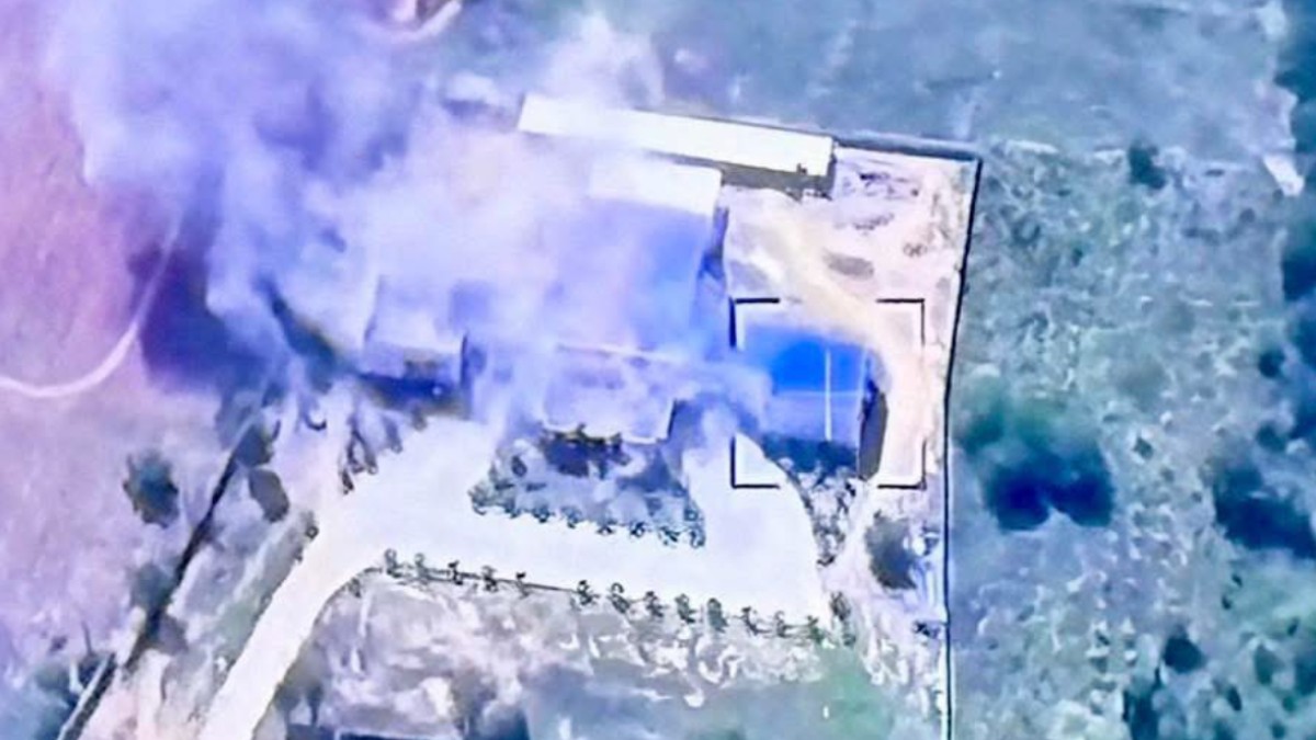 În imagine se vede o filmare aeriană a unei clădiri aflate sub atac. Structura este înconjurată de fum dens, ceea ce sugerează că a fost lovită recent. Zona din jur pare a fi una izolată, cu teren deschis. Î