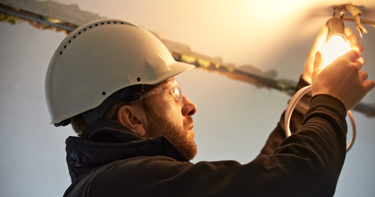 Un electrician cu o cască pe cap care montează un bec și racordează o casă la curent electric