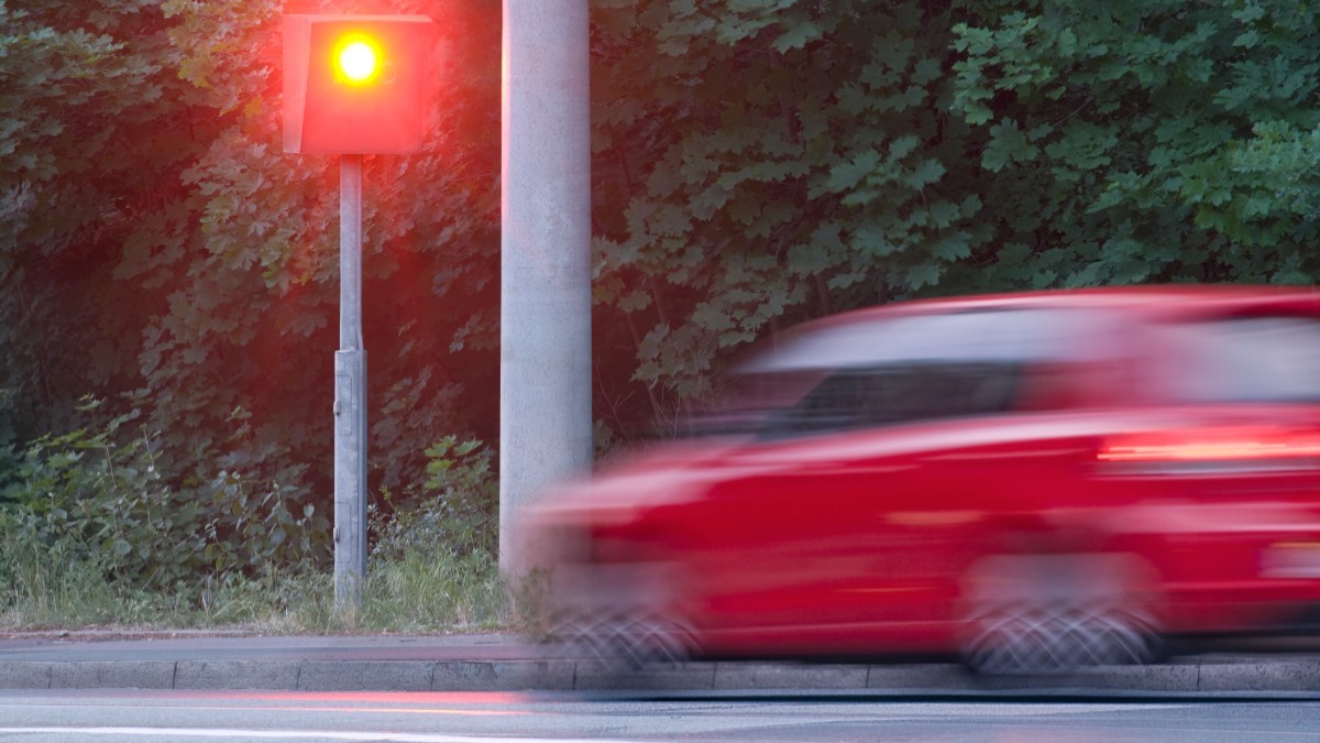 O mașină roșie în mișcare în dreptul unui radar fix de viteză care declanșează un bliț luminos roșu pe marginea drumului.   