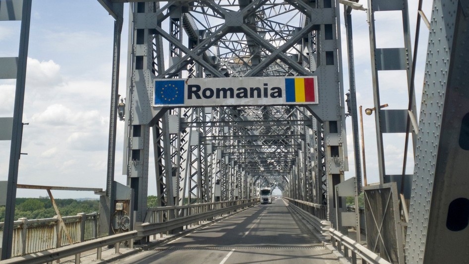 Podul Giurgiu-Ruse, podul Prieteniei, văzut dinspre Giurgiu. Pe șosea se află un TIR alb, iar deasupra ramei metalice ale podului se observă un semn de circulație cu textul „România”, steagul Uniunii Europene și steagul României