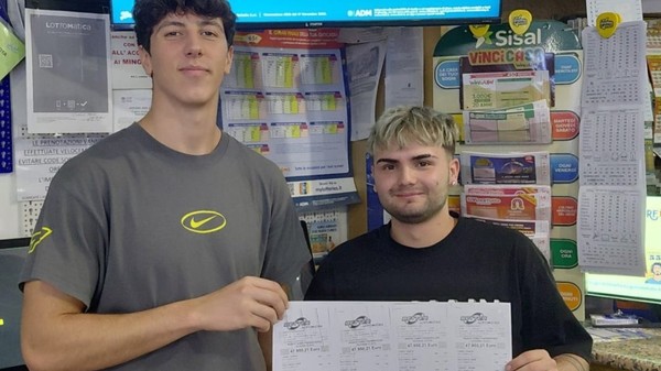 Doi tineri care zâmbesc și țin împreună mai multe bilete de pariu/loterie într-o agenție de jocuri de noroc, pe bilete fiind vizibil un câștig de 47.950,21 euro.