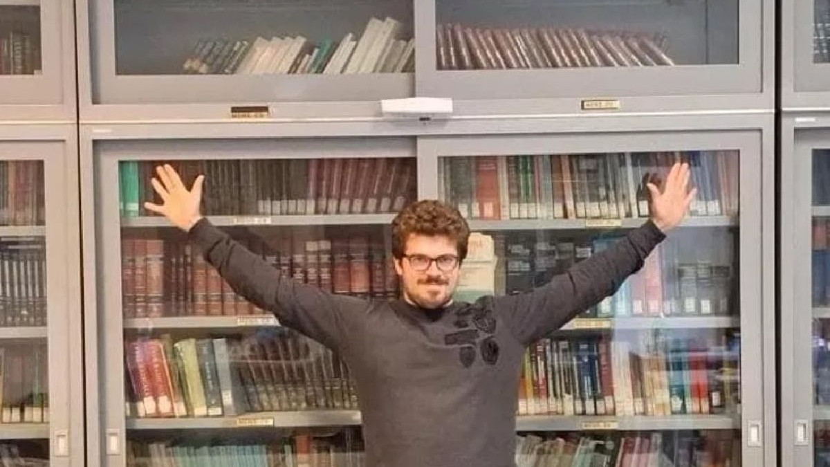Un bărbat cu părul cârlionțat, ochelari și un pulover închis la culoare, cu brațele ridicate, stă în fața unei biblioteci