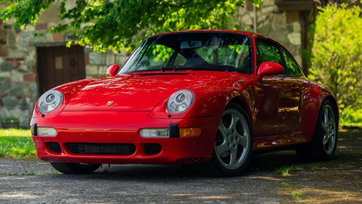 Imaginea prezintă o mașină sport Porsche 911 roșie, parcată pe o alee umbrită de copaci în fața unei clădiri din piatră.