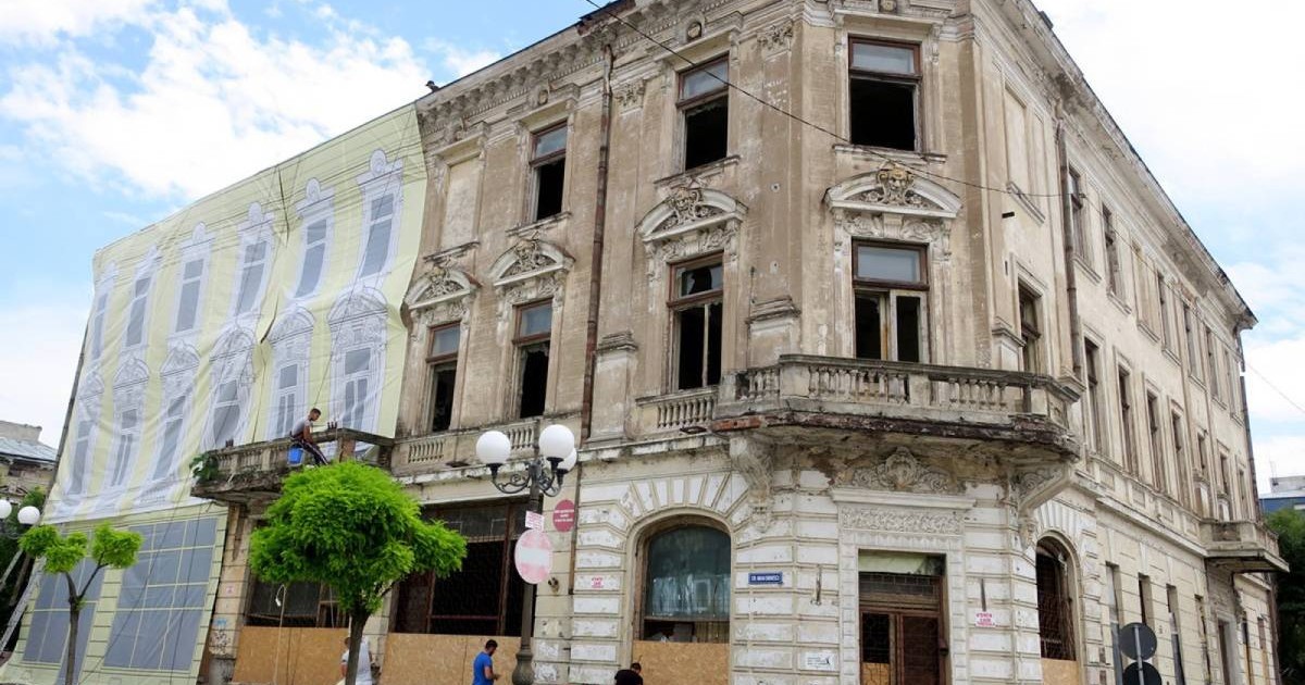 Clădire veche, fostul CEC din Brăila, cu două etaje, foarte degradată, cu fațadă scorojită și ferestre fără geamuri. Balconul este deteriorat, iar intrările de la parter sunt acoperite cu scânduri. Pe partea stângă, imobilul este acoperit cu o plasă imprimată ce imită fațada originală. În față se văd câțiva muncitori, copaci mici și stâlpi de iluminat, iar zona este delimitată cu bandă de siguranță.