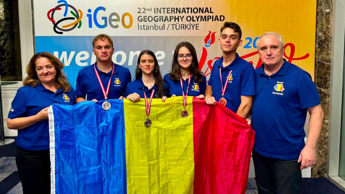 În imagine apar patru elevi români și doi profesori, purtând tricourile lotului României și medaliile obținute la Olimpiada Internațională de Geografie. Echipa ține drapelul României în fața panoului oficial al competiției de la Istanbul.