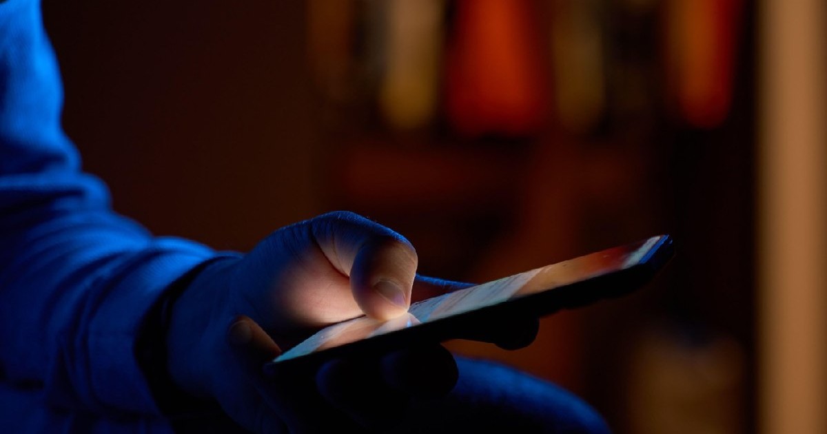 Bărbat folosind smartphone-ul noaptea. Prim-plan cu mâna care ține smartphone-ul, glisând și derulând feed-ul de social media pe ecranul luminos într-o cameră întunecată.
