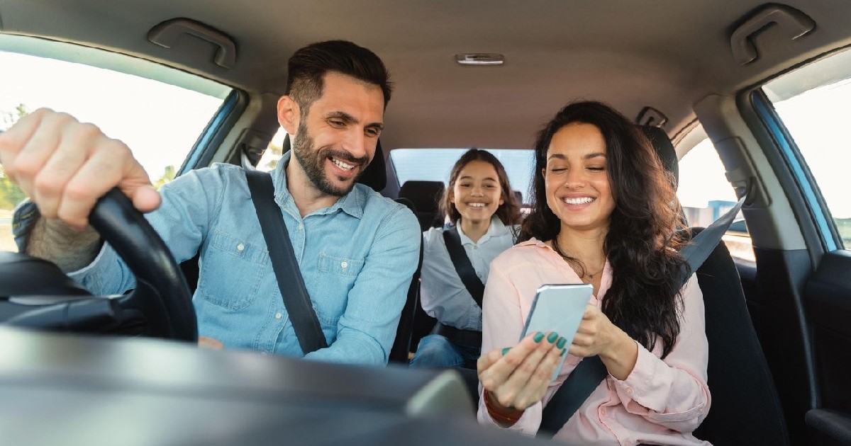 O femeie fericită care arată ecranul telefonului mobil unui bărbat care conduce, pe bancheta din spate este o fetiță care poartă centura de siguranță