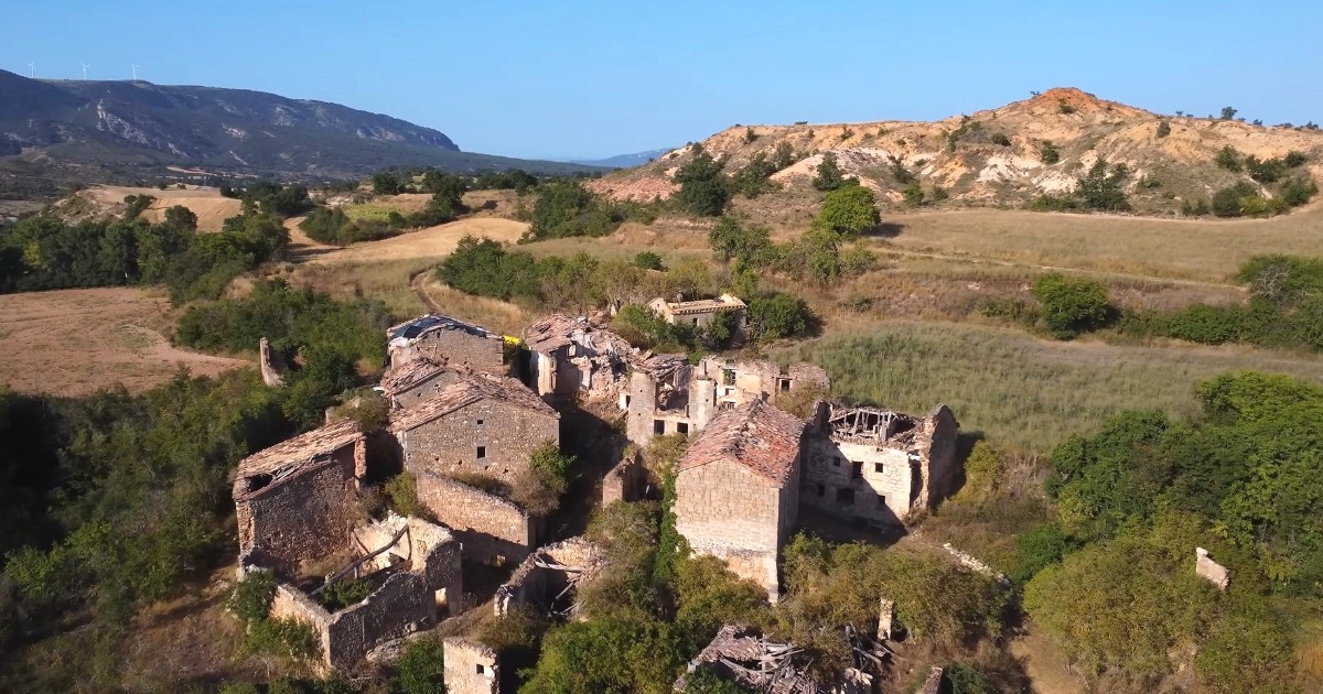 Vedere aeriană a unui sat vechi și părăsit, case din piatră aflate în stare avansată de degradare, cu acoperișurile prăbușite, vegetație sălbatică și dealuri aride sub un cer senin.