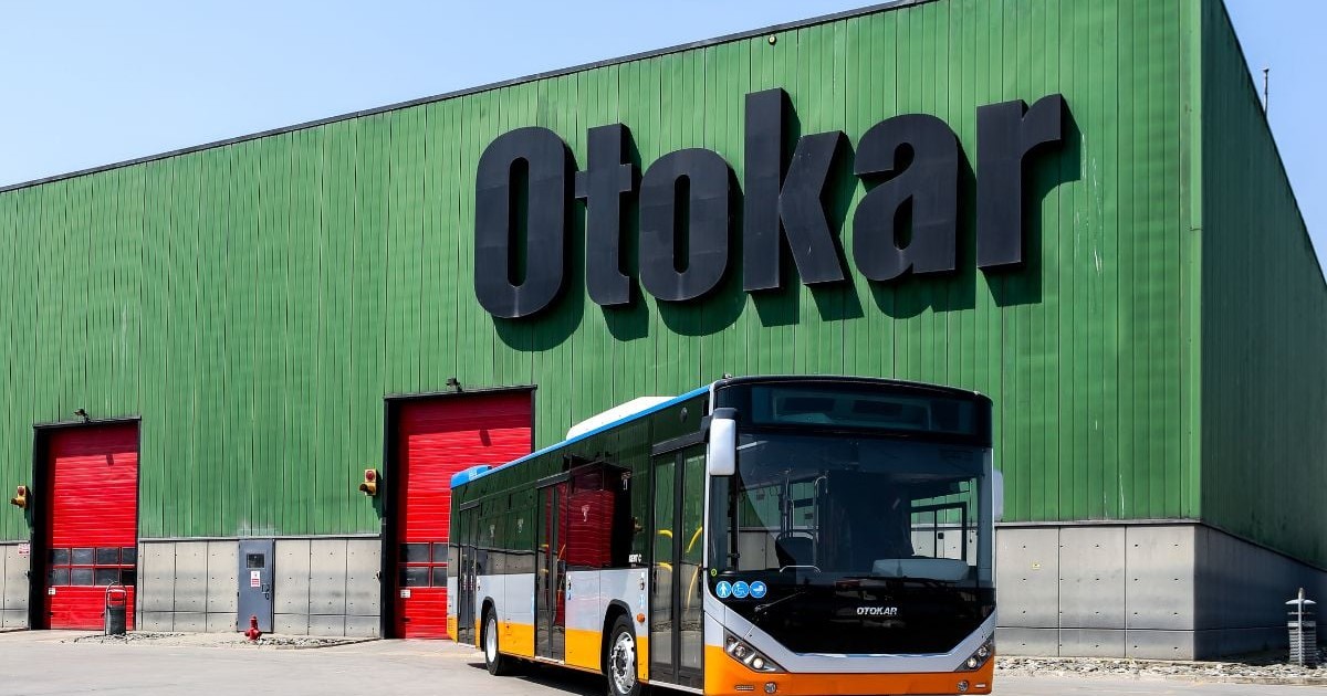 autobuz modern de transport în comun parcat în fața unei clădiri industriale mari, cu pereți din tablă ondulată de culoare verde intens, pe care este scris central, cu litere negre de dimensiuni foarte mari, textul „Otokar”. Autobuzul are o caroserie multicoloră, cu partea inferioară portocalie, o bandă gri la mijloc și geamuri mari, închise la culoare, în timp ce pavilionul prezintă elemente albe și albastre