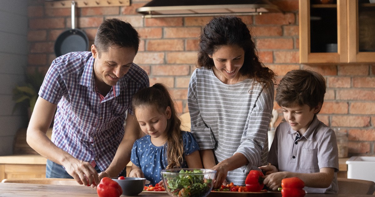 O familie pregătește împreună masa într-o bucătărie modernă, într-o atmosferă caldă și relaxată. Imaginea transmite ideea de prânz gustos, gătit acasă, cu ingrediente simple și accesibile.
