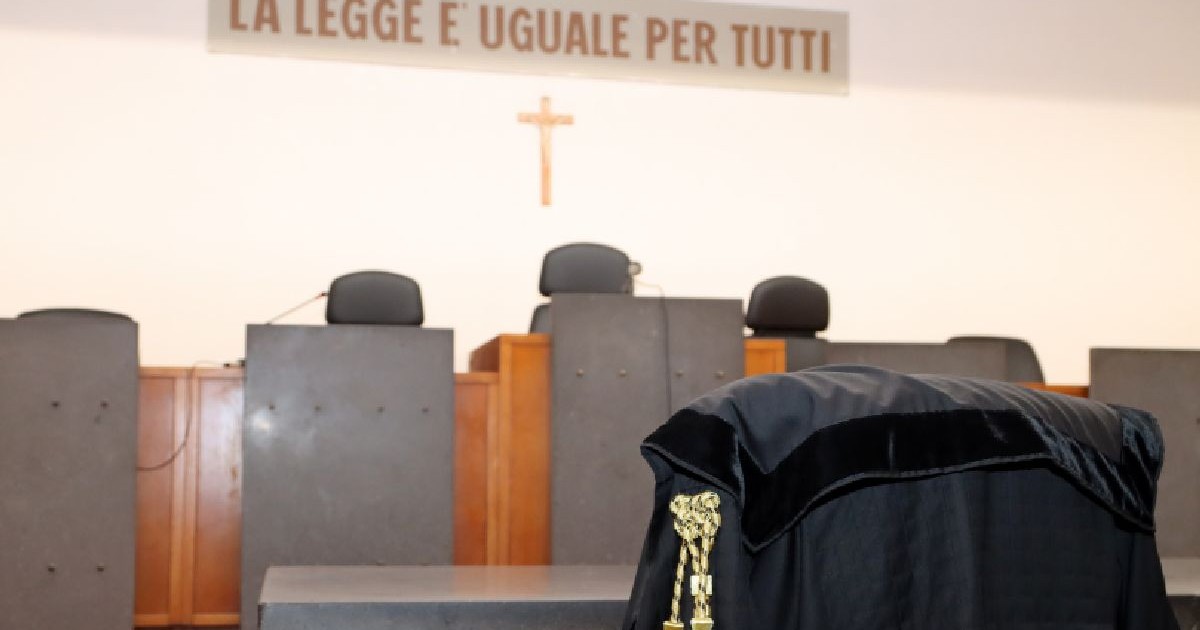 Fotografie a interiorului unei săli de tribunal, cu o robă neagră cu ciucuri aurii aruncată peste un scaun, în fața unei mese din lemn cu mai multe scaune gri, goale, și o cruce pe peretele de deasupra.