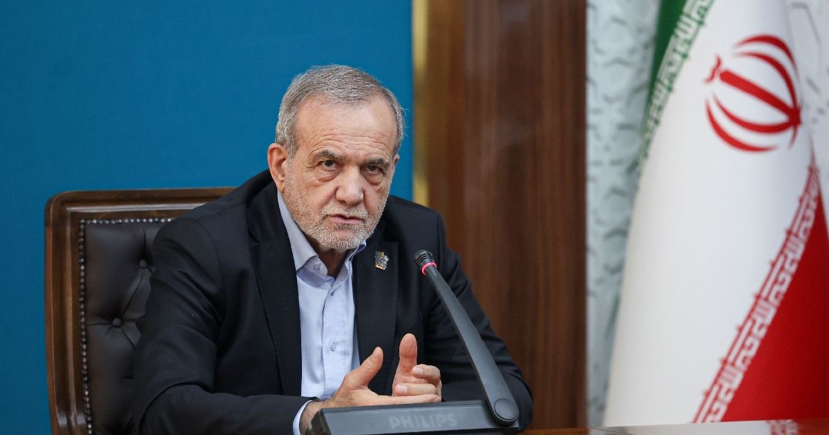 Președintele Iranului, Masoud Pezeshkian, cu părul și barba cărunte, îmbrăcat într-un costum negru și cămașă albastră, așezat la un birou din lemn. În spatele lui se află steagul Iranului.