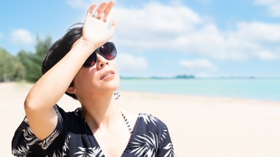 Ce este radiaţia ultravioletă şi cum interpretăm indicele UV  - O femeie brunetă se află pe plajă, la malul mării, într-o ținută ușoară de plajă și cu ochelari, se uită spre soare, ținându-și mână în dreptul ochiilor