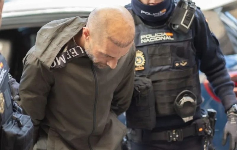 Un bărbat într-o jachetă gri, încătușat, este escortat de doi polițiști