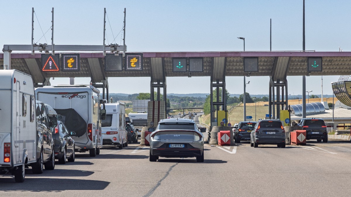 Imaginea prezintă mai multe vehicule, inclusiv rulote și autoturisme, aliniate la benzile unei stații de taxare de pe o autostradă din Franța.