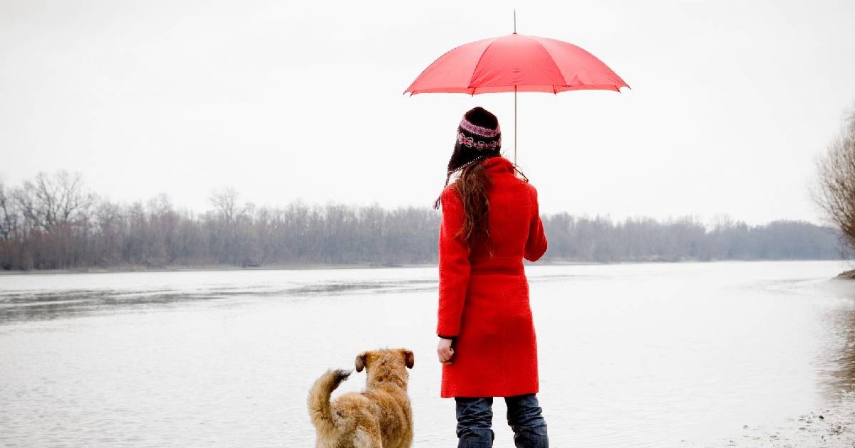 O femeie îmbrăcată cu un palton roșu, ținând în mână o umbrelă roșie și un câine privesc spre un peisaj de toamnă gri