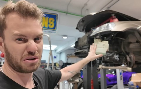 Un bărbat cu o expresie de dezgust, arătând spre zona frontală dezasamblată a unei mașini ridicate pe o platformă într-un atelier auto