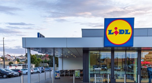 Un magazin Lidl modern este surprins din exterior, cu intrarea largă și logo-ul vizibil deasupra ușilor automate. În fața clădirii se află parcarea și zona pentru cărucioare, sugerând acces facil pentru clienți.