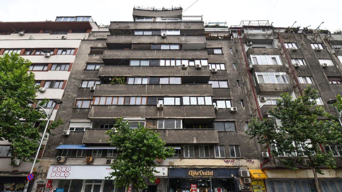 Imaginea prezintă fațada cenușie a unui bloc vechi de locuințe cu mai multe etaje și magazine la parter, situat într-o zonă urbană.