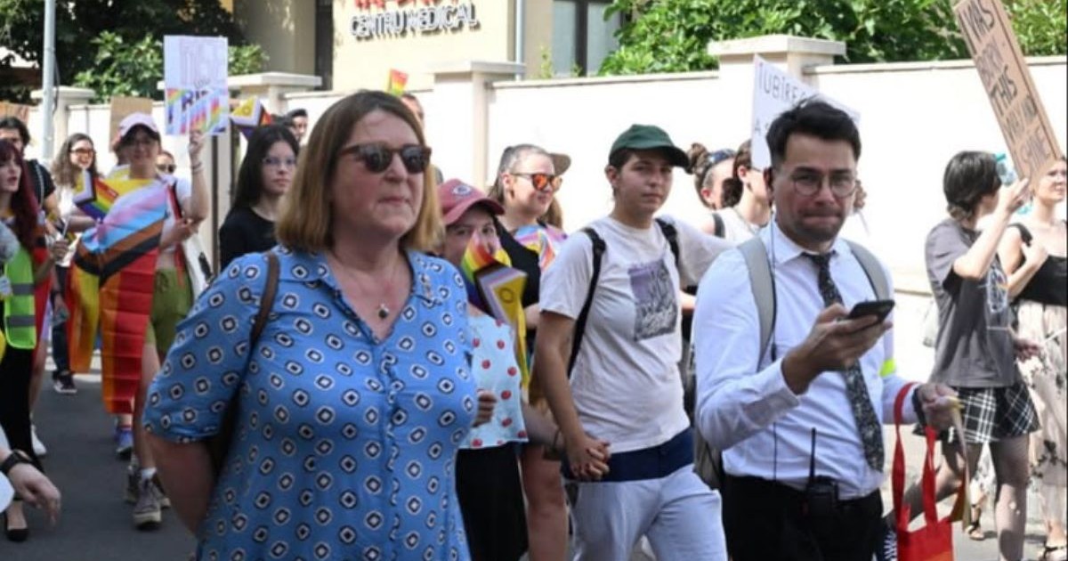 În imagine ambasadoarea Olandei, Willemijn van Haaften, într-o richie bleu, cu ochelari de soare si mainile la spate, participă la Oradea Pride, alături de activistul Victor Ciobotaru, in comasă albă si pantalon negru, in jurul lor mai mult persoane care participa la parada, unele tin pancarde. 26 iulie 2025, Oradea