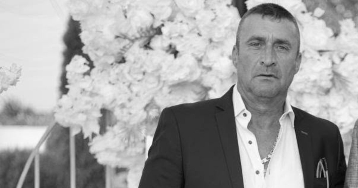 Imaginea îl surprinde pe Radu Bogățeanu îmbrăcat într-un costum închis la culoare și o cămașă deschisă, în fața unui decor festiv realizat din flori albe.