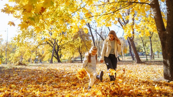O mamă și fiica ei se joacă cu bucurie printre frunzele colorate de toamnă într-un parc însorit, într-o zi senină de octombrie