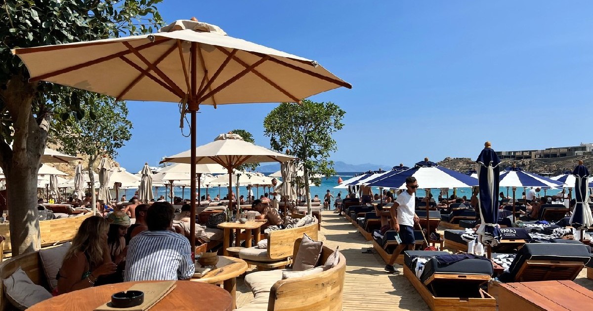Oaspeții se relaxează sub umbrele de soare într-un club de plajă elegant de pe coasta insulei Mykonos, Grecia. Cu scaune elegante, șezlonguri și vedere la Marea Egee.